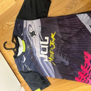 Svart och grå fotbollströja med grafiskt tryck - Säljer en cool fotbollströja från NINETA med svart och grå bas, gula detaljer och grafiskt tryck med texten 'Mise à jour'. Tröjan har korta ärmar och rund hals. Perfekt för dig som vill sticka ut på planen.