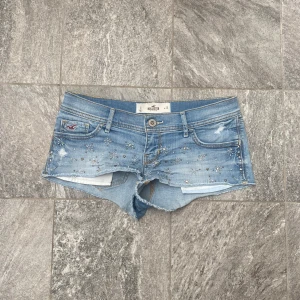 Ljusblå jeansshorts med nitar från Hollister - Lågmidjade  ljusblå jeansshorts från Hollister med slitna detaljer och råa kanter. Shortsen har silverfärgade nitar i stjärnmönster och klassisk femficksdesign. Sjukt trendiga och eftertraktade. Skriv om ni har frågor. Har inga bilder med de på eftersom de inte passar mig ❤️