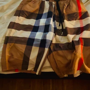 Rutig badshorts från Burberry - Säljer ett par klassiska badshorts från Burberry i storlek XS. Shortsen har det ikoniska rutmönstret i beige, svart, vitt, rött och blått samt snörning i midjan. Perfekta för sommaren och strandhäng!