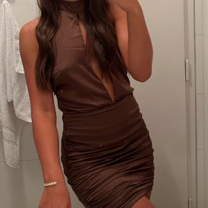 Brun  kortklänning  - Brun tight miniklänning med hög hals och djup slits framtill. Klänningen är ärmlös och har en figurnära passform med lätt rynkade detaljer längs sidorna. 