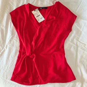 Röd blus från Zara - Säljer en snygg röd blus från Zara med knytdetalj framtill. Toppen har korta ärmar och v-ringning, perfekt för att lyfta vilken outfit som helst. Materialet är lätt och känns skönt mot huden. Helt oanvänd! Lappen kvar💞