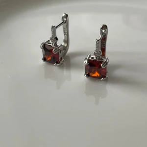Silverörhängen med röd sten 925 - Säljer ett par eleganta örhängen i silver med fyrkantig röd sten och små gnistrande detaljer upptill. Märkta med S925 vilket betyder äkta silver. Perfekta för att ge en färgklick till din look!