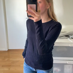 Mörk blå tröja med knappar från H&M - Säljer en stilren mörk blå tröja från H&M i storlek M. Tröjan ganska liten i storlek då jag brukar ha XS/S. Tröjan har långa ärmar och dekorativa knappar på axeln som ger en extra touch. Perfekt för en enkel och elegant look.