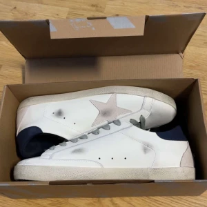 Vita sneakers från Golden Goose - Säljer ett par vita sneakers från Golden Goose med grå snörning, ljusgrå stjärndetalj på sidan och mörkblå häl. Skorna har en klassisk låg modell och texten GGDB/SSTAR på sidan. Perfekta för en avslappnad och trendig stil.