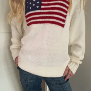 Vit stickad tröja med amerikansk flagga - Vit stickad tröja med en stor amerikansk flagga i rött, vitt och blått på bröstet. Tröjan har lång ärm och en klassisk, avslappnad passform. Perfekt för dig som vill sticka ut med en cool detalj på vardagen.