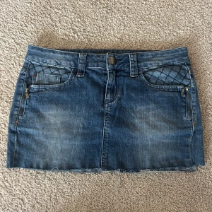 Lågmidjad jeanskjol  - Säljer en blå jeanskjol med snygga quiltade detaljer på fickorna framtill och rå kant nertill. Kjolen har dragkedjor på fickorna och klassisk knappstängning. Perfekt för dig som gillar en cool och avslappnad stil.