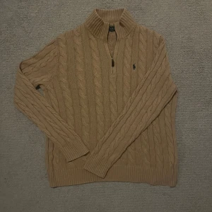  Polo Ralph Lauren zip  - Säljer en beige stickad tröja från Polo Ralph Lauren med klassiskt kabelmönster och half zip framtill. Tröjan har långärmad design, ribbad krage och muddar. Perfekt för dig som gillar stilrena och tidlösa plagg med snygg passform.