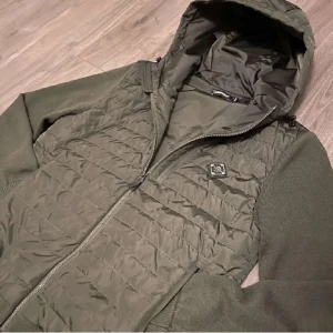 Cardigan från J.Lindeberg - Snygg olivgrön pufferjacka från J.Lindeberg med quiltad front och huva. Jackan har stickade ärmar och muddar, dragkedja framtill och en diskret logga på bröstet. Perfekt för dig som vill ha en stilren och sportig look.