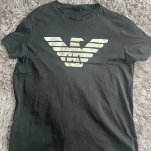 Svart armani t-shirt - Svart t-shirt från Armani med stor vit örnlogga och GA-tryck på bröstet. Klassisk rund hals och korta ärmar. Storlek M men sitter som S. Små hål på fram och baksida.