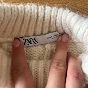 Vit stickad väst från Zara - Säljer en vit stickad väst från Zara med grov kabelstickning och ribbad halsringning. Västen är ärmlös och har en boxig, croppad passform som ger en trendig look. Perfekt att slänga över en skjorta eller långärmad tröja för extra stilpoäng.