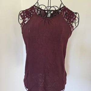 Mörklila ärmlös topp med spets - Supersnygg mörklila ärmlös topp med rund halsringning och detaljerade spetsinslag på axlarna. Toppen är tillverkad i viskos med spets i polyester, vilket gör den både mjuk och luftig. Perfekt för dig som gillar en feminin och stilren look.