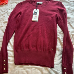 Vinröd stickad tröja från Lindex - Snygg vinröd stickad tröja från Lindex med rund hals och långa ärmar. Tröjan har dekorativa knappar i guld på ärmsluten och en liten metallbricka vid nederkanten. Perfekt för dig som gillar stilrena detaljer och skön passform.