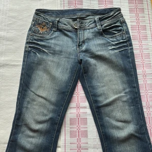  bootcut jeans med broderi - Snygga blå bootcut jeans från K-Star med coolt broderade fickor och pärldetaljer både fram och bak. Jeansen har låg midja, klassisk dragkedja och slitningar i jeans materialet