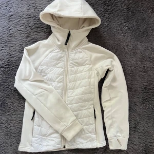 Cremevit hybridjacka från Didriksons Tovik  - Snygg vit höstjacka från Didriksons med huva och hel dragkedja framtill. Jackan har quiltade paneler på framsidan, två sidofickor med dragkedja och långa ärmar med tumhål. Perfekt för dig som gillar stilren och sportig look. Tovik Didriksson går även att ha som ett lager under en skaljacka 