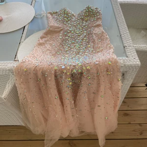 Champagnefärgad långklänning med paljetter - Säljer en magisk champagnefärgad långklänning med glittriga paljetter och stenar över hela klänningen. Klänningen är axelbandslös med en hjärtformad urringning och har ett svepande tylltyg som ger extra volym. Perfekt för bal eller fest! Dragkedja baktill för enkel påklädning.