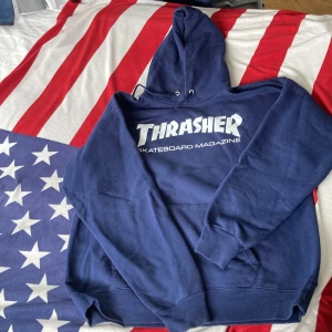 Oanvänd Mörkblå Thrasher hoodie med tryck - Säljer en mörkblå hoodie från Thrasher med stort vitt tryck på bröstet. Tröjan har huva med snörning och en klassisk känguruficka framtill. Perfekt för dig som gillar streetwear och skate. Materialet är mjukt och känns skönt mot huden.