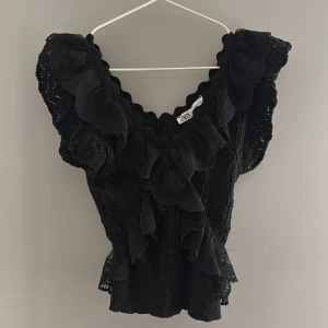 Svart volangblus från Zara - Supersnygg svart blus från Zara med volanger och spetsdetaljer. Djup v-ringning och korta ärmar med extra volym. Perfekt för dig som gillar romantisk och trendig stil. Blusen är i ett stickat spetsmaterial som känns både luftig och cool.