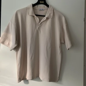 Beige kortärmad pikétröja från Zara - Snygg beige pikétröja från Zara i storlek L. Tröjan har klassisk krage, två knappar vid halsen och är tillverkad i mjuk bomull. Passformen är loose och den är perfekt för dig som gillar enkel och clean stil. Färgen är neutral och funkar till många outfits.