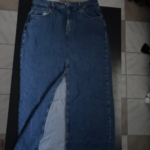 Blå lång jeanskjol med slits - Säljer en klassisk blå lång jeanskjol från Bikbok med låg midja, fem fickor och en snygg slits framtill. Kjolen har bälteshällor och stängs med dragkedja och knapp.