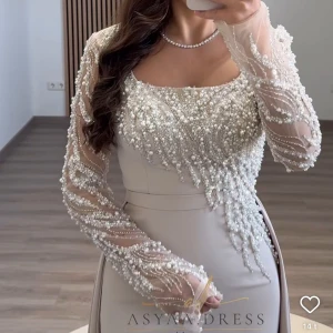 Champagnefärgad långklänning med pärlor - Säljer en elegant långklänning i champagnefärg från Asyaa Dress. Klänningen har långärmade meshärmar dekorerade med pärlor och paljetter, samt en figurnära passform med slits. Perfekt för dig som vill sticka ut med lyxiga detaljer och unik design. Använd 1 gång på min förlovning. Ursprungspris 4000, mitt pris 3000. 