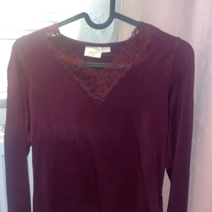 Vinröd långärmad topp med spets - Snygg vinröd långärmad topp med v-ringad hals och dekorativ spetsdetalj framtill. Mjuk och skön känsla, perfekt att styla med jeans eller kjol. Färgen är djup och mörk, och spetsen ger en extra touch av stil.