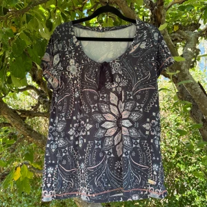 Odd Molly blommig topp med knyt - Supersnygg träningstopp från Odd Molly med blommigt och paisley-inspirerat mönster i mörkblått, rosa och vitt. Rund halsringning med knyt framtill, korta ärmar och mjukt bomullsmaterial. Perfekt för dig som gillar detaljer och coola prints. Storlek 3 som enligt deras storleksguide motsvarar en storlek L. Men kan vara något liten i storlek.  Mycket fint skick, men kan vara något blekare än bilder visar. 