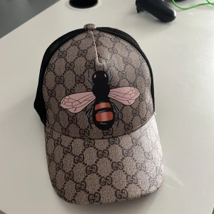  keps med bi och GG-mönster - Säljer en snygg keps med klassiskt GG-mönster i beige och brunt, stor bi-print framtill och svart mesh på sidorna och bak. Justerbar rem med grönt och rött band bak. Perfekt för dig som vill sticka ut med designerstil.