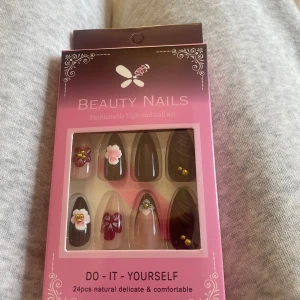Beauty Nails konstgjorda naglar med blommor - Säljer ett set med 24 konstgjorda naglar i olika storlekar. Naglarna har en elegant spetsig form och kommer i väldig mörk röd, nästan svart och nude nyanser med detaljer  och små guldfärgade pärlor. Ingår en fil och klister 