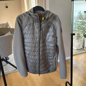 Cardigan från Parajumpers - Nypris ligger omkring: 4500 kr. Snygg beige cardigan från Parajumpers med quiltad framsida och mesh-liknande ärmar. Jackan har huva, gul detalj vid halsen och dragkedja framtill samt bröstficka med zip. Märkespatch på vänster arm. 