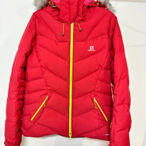 Röd dunjacka från Salomon, storlek L - Riktigt snygg röd dunjacka från Salomon med limegröna dragkedjor och huva med fuskpäls. Jackan har två fickor framtill, justerbar huva och coolt mönstrad sömnad. Perfekt för kalla dagar när du vill sticka ut lite extra.
