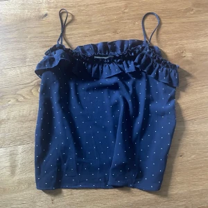 Mörkblå prickig linnetopp med volang - Supersöt mörkblå linnetopp med tunna axelband och små vita prickar. Toppen har en volang längs urringningen. Perfekt för varma dagar när du vill ha något lätt och luftigt på dig.