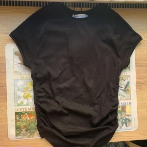 Svart croppad topp från Bershka - Svart croppad topp från Bershka i mjuk bomull. Modellen har korta ärmar och snygg rynkning på sidorna som ger en cool siluett. Perfekt att styla med jeans eller kjol för en chill och trendig look. Prislappen är kvar på