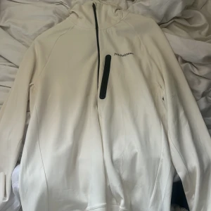 Vit hoodie Peak Performance med half zip - Säljer en stilren vit hoodie från Peak Performance i storlek M. Tröjan har huva, svart half zip och en praktisk bröstficka med dragkedja. Materialet är mjukt och känns som polyester, perfekt för kyliga dagar. Loggan är diskret tryckt på bröstet.