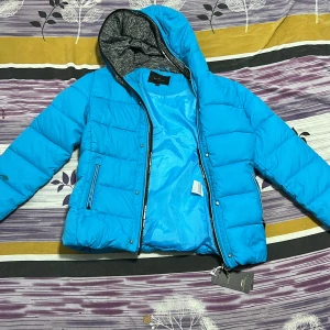 Blå pufferjacka från Lanter XL - Säljer en blå pufferjacka från Lanter i storlek XL. Jackan har huva med gråmelerat foder, två sidofickor med dragkedja och quiltad design. Materialet är syntet och jackan stängs med dragkedja framtill. Perfekt för kalla dagar när du vill ha en chill och sportig look. Är i bra skick, men har en gul fläck 