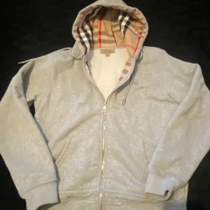 Grå Burberry hoodie med dragkedja - Säljer en grå hoodie från Burberry med klassiskt rutigt mönster i huvan. Tröjan har dragkedja framtill, snörning vid huvan och två fickor. Perfekt för dig som gillar stilrena märkesplagg med lyxig touch och snygga detaljer.