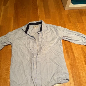 Ljusblå skjorta Hampton Republic - Snygg ljusblå skjorta från Hampton Republic med mörkblå krage och broderad fågel på bröstet. Klassisk knappstängning framtill och långärmad modell. Perfekt för dig som gillar stilrena och enkla plagg med en liten detalj.