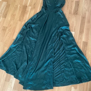 Mörkgrön långklänning från Fashion Nova - Säljer en elegant mörkgrön långklänning från Fashion Nova med glansigt tyg och axelband. Klänningen har en figurnära överdel och ett svepande, utsvängt kjolparti. Materialet är mjukt och har en satinliknande finish. Dragkedja i sidan för enkel påklädning.