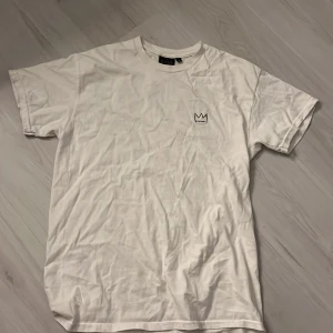Vit Hov1 tour t-shirt med tryck - Vit t-shirt från Hov1 med liten krona broderad på bröstet och stort tour-tryck på ryggen med turnédatum och städer. Klassisk passform och rund hals. Perfekt för dig som gillar musik och vill ha en snygg merch-tröja.