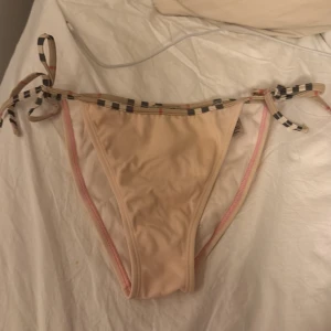 Ljus rosa  bikiniunderdel från Burberry - Snygg ljus rosa  bikiniunderdel från Burberry med klassiskt rutigt mönster i svart, vitt och rött på banden. Modellen har knyt i sidorna för perfekt passform och är tillverkad i mjuk polyamid med elastan. Perfekt för strandhäng och poolparty!