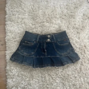 Blå jeanskjol med volangkant - Säljer en blå jeanskjol med volangkant och dubbla knappar framtill. Kjolen har klassiska fickor både fram och bak, med snygga broderade detaljer på bakfickorna. Perfekt för dig som gillar en cool och avslappnad stil.
