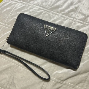 Svart plånbok från Guess med logga - Snygg svart plånbok från Guess med diskret mönster och metalltriangel med logga framtill. Plånboken har dragkedja runtom och en praktisk handledsrem. Materialet är skinnliknande och ger en lyxig känsla. Perfekt för att hålla koll på kort och cash. Som ni ser på tredje bilden har den blivit skrapad och har en skråma men är inte använd.