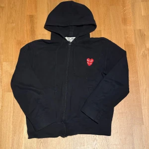 Svart hoodie Comme des Garçons Play - Svart hoodie från Comme des Garçons Play med dragkedja och huva utan defekter. På bröstet finns det ikoniska röda hjärta med ögon broderat. Tröjan är tillverkad i mjuk bomull och har långa ärmar. Perfekt för dig som gillar streetwear och vill sticka ut med en cool detalj.