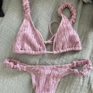 Ljusrosa bikini  - Supersöt ljusrosa bikini med volangdetaljer och ribbad struktur. Bikinin har en klassisk trekantsmodell på toppen med scrunchade axelband och matchande scrunchade sidor på underdelen. 