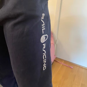 Mörkblå mjukisbyxor Sail Racing - Mörkblå mjukisbyxor från Sail Racing med logga längs benet. Byxorna har resår i midjan och är tillverkade i en mjuk bomulls- och polyestermix. Perfekta för chill dagar eller när du vill ha en sportig look.