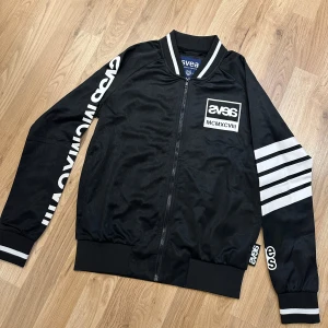 Svart bomberjacka från Svea XS - Svart bomberjacka från Svea med vita detaljer och tryck på ärmarna. Jackan har dragkedja framtill, ribbade muddar och en cool logga på bröstet. Materialet är polyester och jackan har en sportig vibe med vita ränder på ena ärmen. Helt oanvänd men fått en liten fläck bak på ryggen.