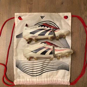 Adidas Predator fotbollsskor strl 38 - Säljer ett par Adidas Predator Elite fotbollsskor i storlek 38. Skorna är vita med svarta och röda detaljer, har snörning och en platt sula med dobbar för gräsplan. Ovandelen är tillverkad i syntetmaterial och designen är riktigt sportig med Predator-logga på sulan och tungan.
