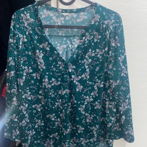 Grön blommig blus med v-ringning från HM  - Säljer en grön blus med v-ringning och vita blommor över hela tyget. Blusen har långa ärmar och knappar framtill. Perfekt för dig som gillar mjuka material och ett avslappnat men ändå snyggt uttryck. Passar till jeans eller kjol för en chill look.