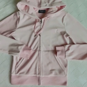 Snygg Ljusrosa Juicy Couture hoodie XS nyskick - Säljer en ljusrosa hoodie från Juicy Couture i storlek XS. Använd väldigt få gånger och är i nyskick. Tröjan har huva med snörning, dragkedja med metall-J och broderad jc logga på bröstet. Mjukt velour material och ribbade muddar vid ärmar och nederkant. 