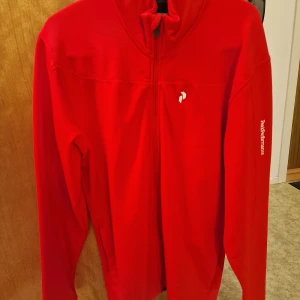 Oanvänd Röd Peak Performance fleece XL - Säljer en snygg röd fleece tröja från Peak Performance i storlek XL. Tröjan har hel dragkedja framtill, lång ärm och hög krage. Loggan finns på bröstet och Peak Performance-tryck på ärmen. Perfekt för dig som gillar sportig och clean stil. Rekommenderas starkt för kallare väder.