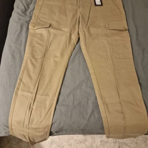 Beige cargo pants Peak Performance  - Säljer ett par snygga beige cargo pants från Peak Performance, modell Aaron. Byxorna har flera praktiska fickor med lock och knapp, klassisk rak passform och är tillverkade i ett slitstarkt bomullsmaterial. Perfekt för dig som gillar streetwear, vandring och funktionella detaljer.
